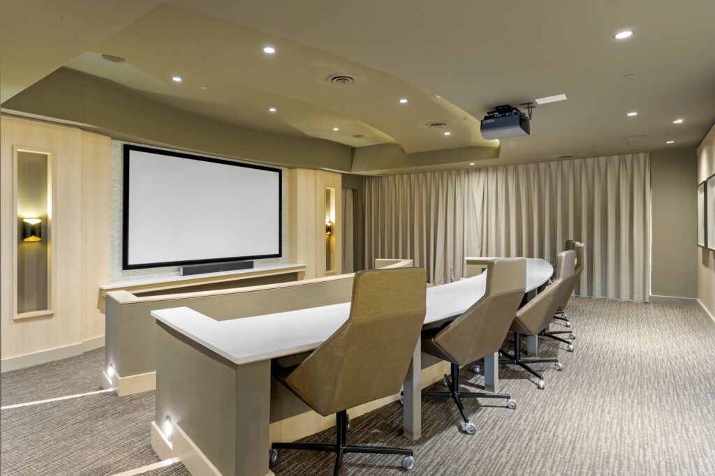 Presentation room with large projection screen, designer lighting, desks with chair seating, and carpet flooring