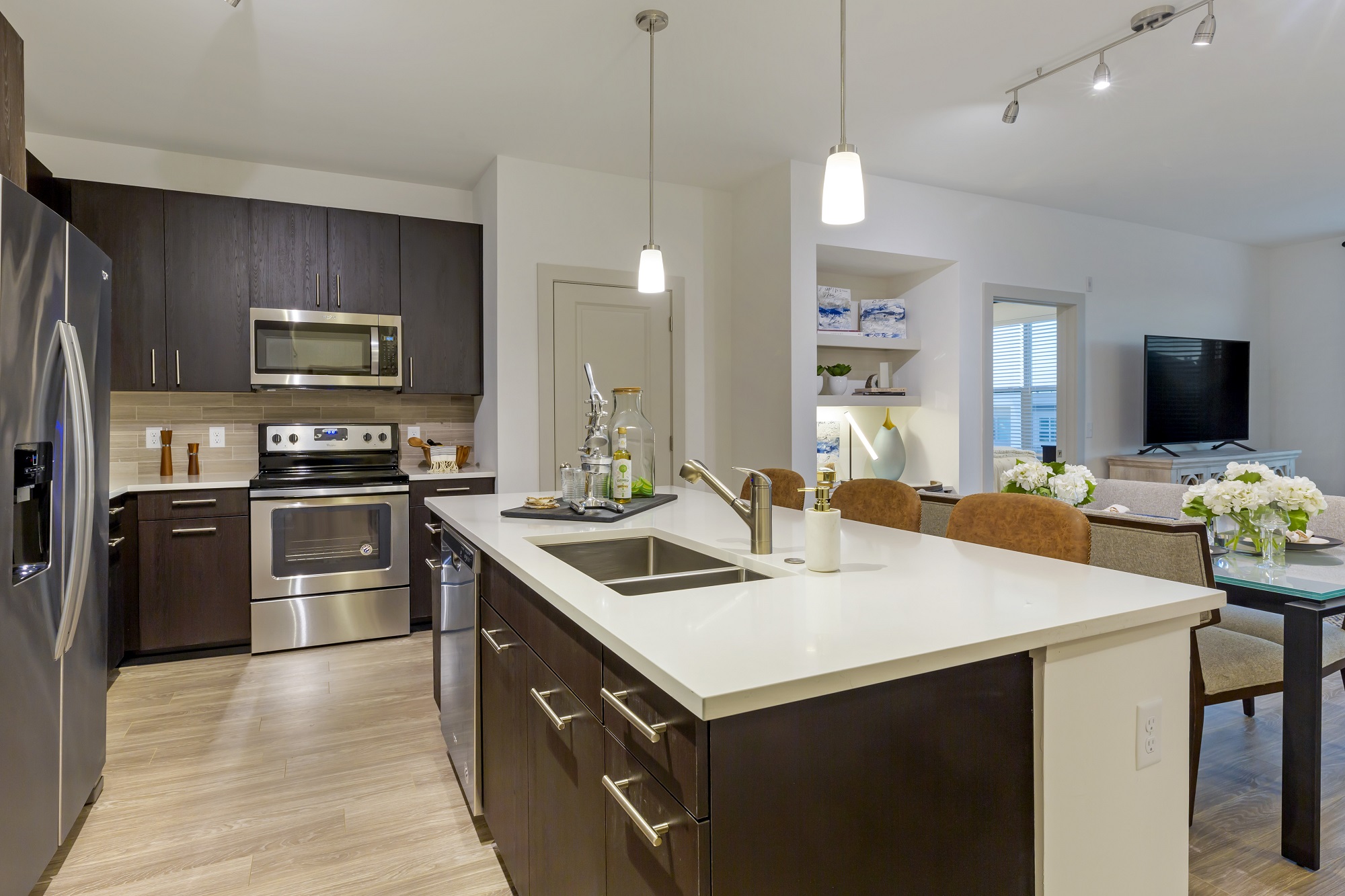 Kitchen/dining/living area with stainless steel appliances, island with sink and bar seating, wood like floor and designer lighting